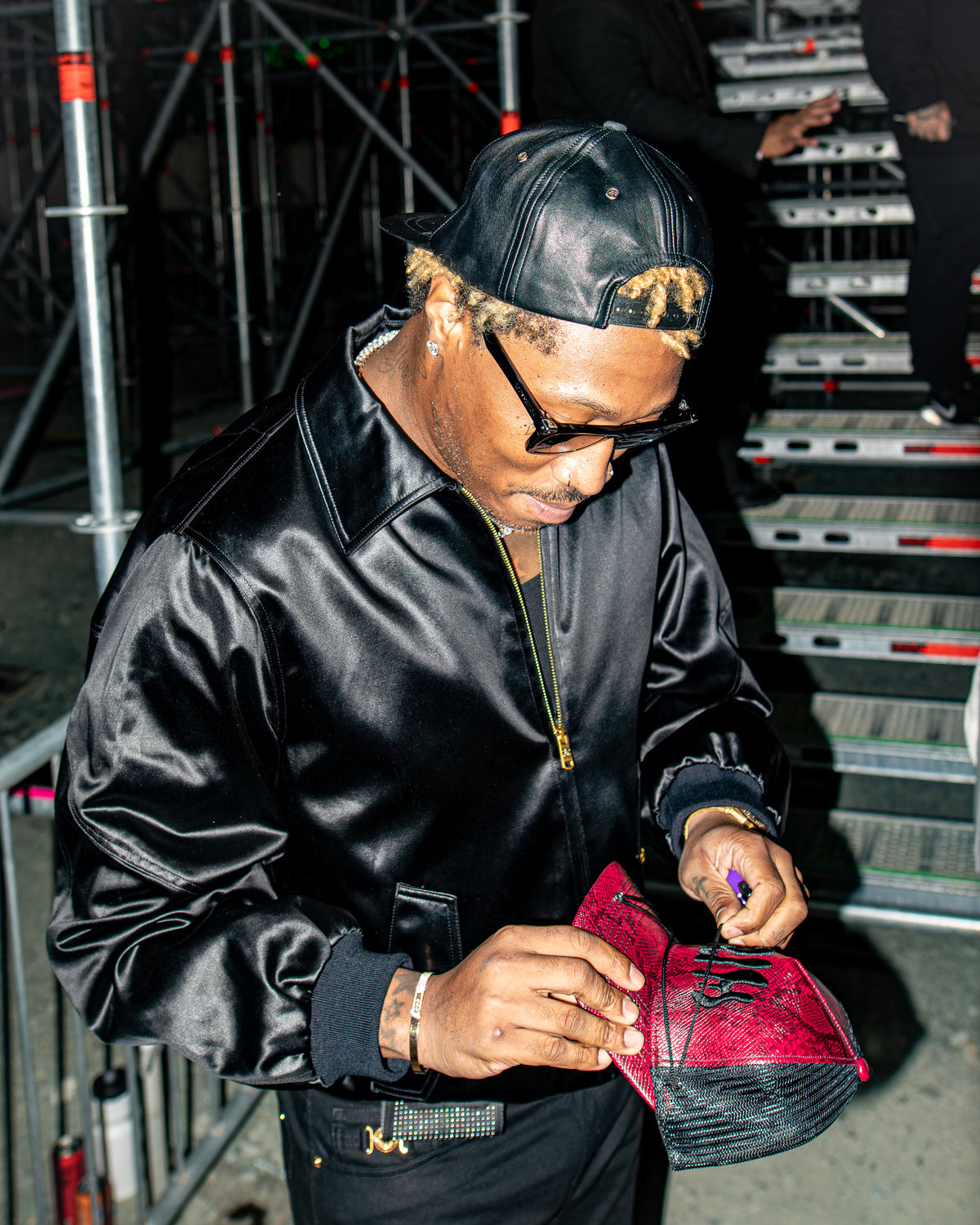 A man in a black satin bomber jacket, black cap, and glasses signs a red and black sneaker backstage on metal stairs. FUTURE DS2 WORE MY CLOTHING BRAND GRIM