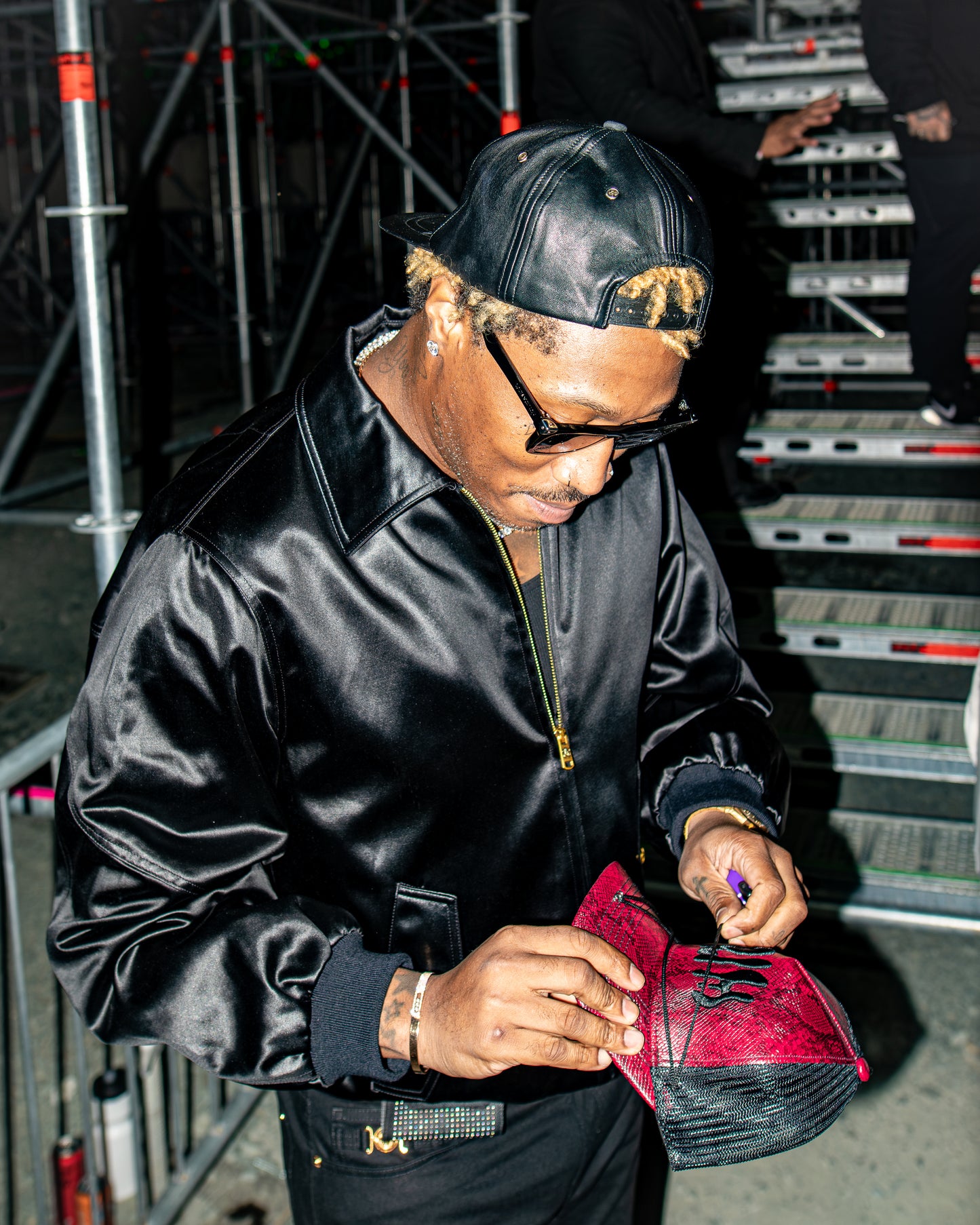 A man in a black satin bomber jacket, black cap, and glasses signs a red and black sneaker backstage on metal stairs. FUTURE DS2 WORE MY CLOTHING BRAND GRIM